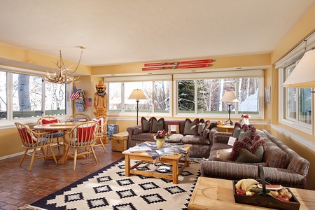 A cozy living room features a dining area with antler chandelier, large windows, sofas with red pillows, wooden coffee table, and ski decor. A snow-covered view is visible outside.