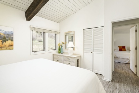 A white bedroom with a bed, dresser with a lamp and flowers, and a window. A second room with a bed is visible through an open door. There is wooden flooring and a beam on the ceiling.