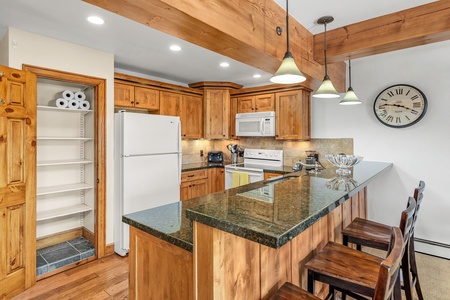 A kitchen with wooden cabinets, granite countertops, a white refrigerator, stove, and microwave. There is a pantry with rolls of paper towels, bar stools by the counter, and a large clock on the wall.