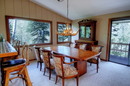 A dining room features a large wooden table, six upholstered chairs, and an antler chandelier. Large windows and a door offer views of surrounding trees, while a bar with stools is in the corner.