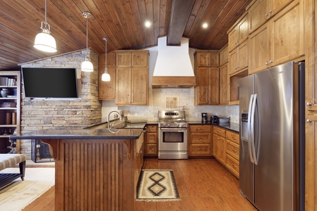 A modern kitchen featuring wooden cabinets, a stainless steel refrigerator and oven, a central island with a sink, pendant lights, a wall-mounted TV, and a cozy stone accent wall.