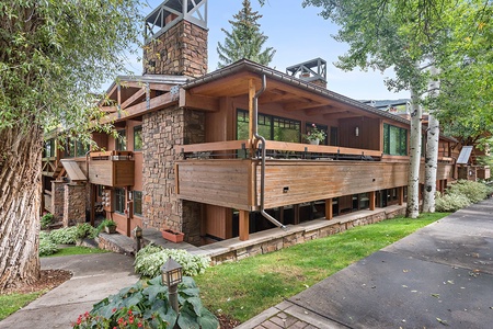 A two-story building with wood and stone exterior, featuring balconies, large windows, and surrounded by greenery, including trees and shrubs. A pathway runs alongside the building.