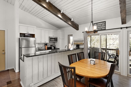 A modern kitchen with white cabinets, stainless steel appliances, and a breakfast bar. Adjacent is a round wooden dining table with six chairs. Large windows offer a view outside.