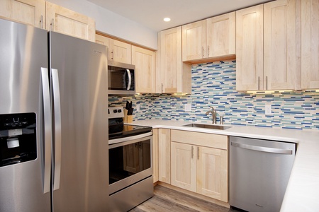 Modern kitchen with stainless steel appliances, light wood cabinets, a tiled backsplash in shades of blue and white, and a wood flooring. Features a sink, oven, microwave, refrigerator, and dishwasher.