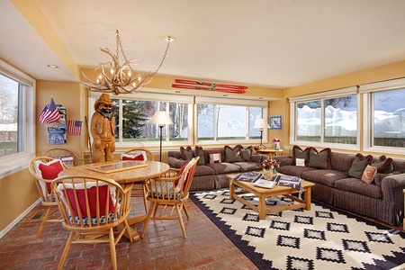 A cozy living room with large windows, a sectional sofa, a wooden dining table set, and a rustic decor featuring a deer antler chandelier, wooden bear statue, and red ski decor on the wall.