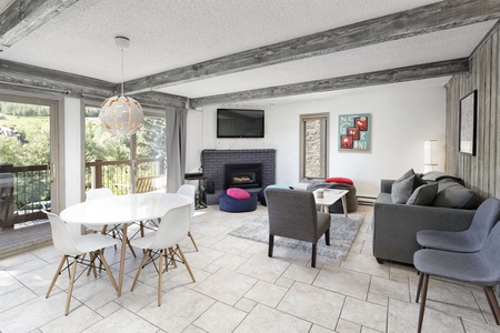 A modern living room with a dining area, featuring a white table with chairs, a gray sofa, a fireplace, and large windows overlooking a green landscape.