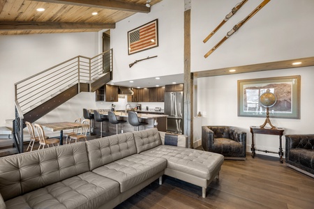 Modern living room featuring a gray sectional sofa, open kitchen with island and bar stools, upstairs loft, American flag and skis on the wall, rustic dining table with metal chairs, and a globe.