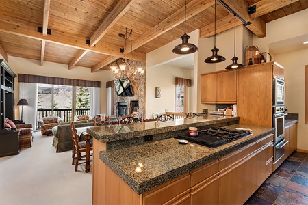 A spacious kitchen with granite countertops opens to a living area featuring large windows, a stone fireplace, wood-beamed ceilings, and a mix of comfortable seating, with views of a mountainous landscape.