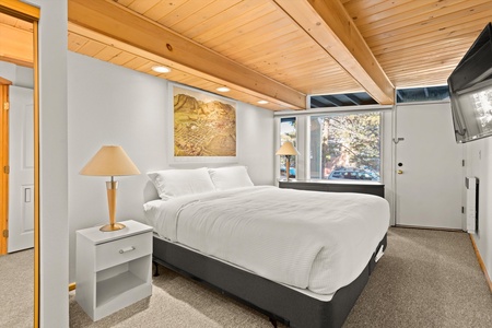 A cozy bedroom with a white bed, two bedside tables with lamps, a wall-mounted TV, a window with a view of trees, and a wooden ceiling with exposed beams.