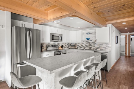 Modern kitchen with white cabinets, stainless steel appliances, marble countertop island with bar stools, wood-paneled ceiling, and a mosaic tile backsplash.