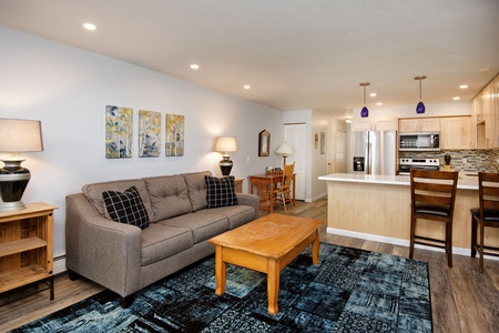 A modern open-plan living room and kitchen with a gray sofa, wooden coffee table, blue area rug, and a dining table. The kitchen features light wood cabinetry, stainless steel appliances, and a breakfast bar.