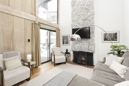 A modern living room with a stone fireplace, flat screen TV, large windows, beige furniture, and wooden accents. A floor lamp arches over a gray sofa. Snow is visible outside through a sliding door.