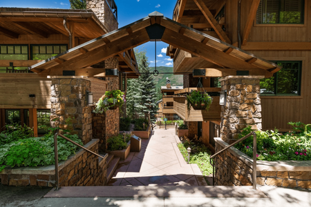 Wooden walkway with stone pillars and hanging plants leads to a scenic view of rustic buildings and trees.