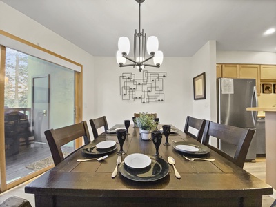 A dining table set for six with black plates, gold utensils, and black goblets is in a room with a modern chandelier, art on the wall, a sliding glass door, and a view into a kitchen with wooden cabinetry.