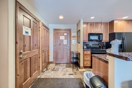 A kitchen area and hallway with wood cabinets and doors. The kitchen includes a stove, microwave, and countertops. Various signs are posted on the doors.