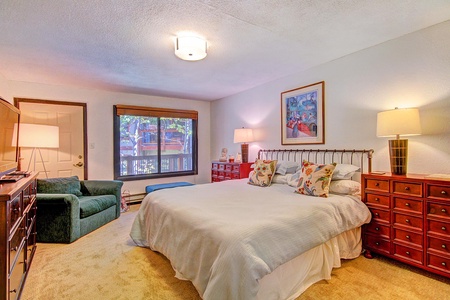 A bedroom with a double bed, two wooden nightstands with lamps, a green armchair, a window, and wall art. The carpet is beige, and the walls are light-colored. Natural light filters through the window.