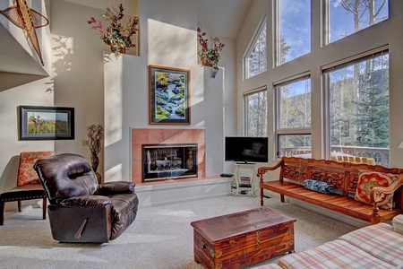 A living room with large windows, a fireplace, a recliner, wooden bench, ornate trunk, TV, and wall art. Sunlight streams in, illuminating the carpeted floor and furniture.