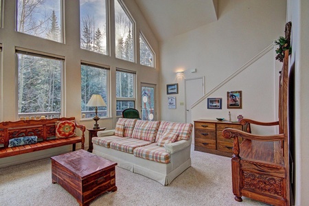 A living room with a high ceiling, large windows, and forest views. There is a plaid sofa, wooden coffee table, armchairs, wall art, and a staircase leading upstairs.