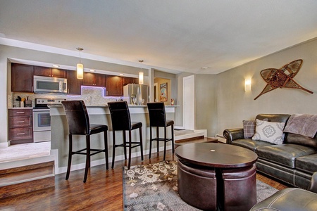 A modern open-plan living room and kitchen with dark wood cabinetry, a three-stool breakfast bar, dark leather seating, and decorative snowshoes on the wall.