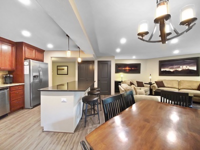 Spacious kitchen and living area with wooden flooring, an island with stools, stainless steel appliances, dark wood cabinets, a dining table, and a sectional sofa with wall art and lighting fixtures.