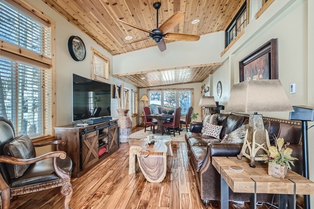 A cozy living room with wooden ceiling and floors, featuring a leather sofa, armchairs, a large wall-mounted TV, and a ceiling fan. A dining area with a table and chairs is in the background.