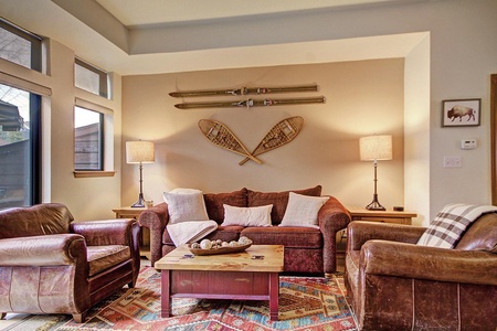 A cozy living room with a brown sofa, two leather armchairs, a wooden coffee table, and rustic wall decor featuring vintage skis and snowshoes. Two lamps flank the sofa on side tables.