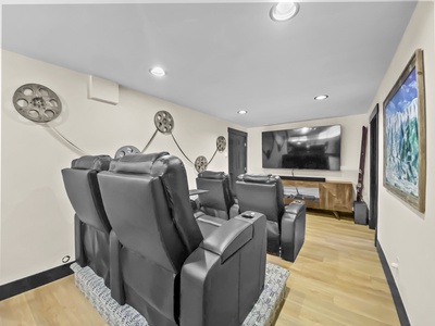 A home theater with four black leather recliners facing a large flat-screen TV on a wall. Old film reels are mounted on the wall as decoration. A framed painting is on the right wall.