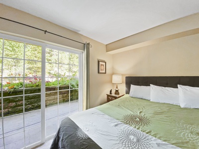 A bedroom with a large bed featuring a green and white bedspread, a bedside table with a lamp, and sliding glass doors leading to an outside garden area.