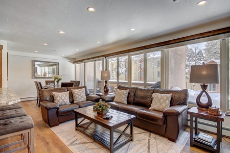 A modern living room features brown leather sofas, a wooden coffee table, a dining area with a decorative mirror, and large windows letting in ample natural light.