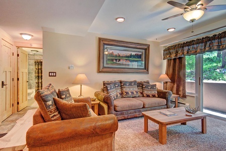 A cozy living room featuring a brown couch and armchair with bear-themed pillows, a wooden coffee table, a landscape painting above the couch, and a ceiling fan. Decor includes lamps and large windows.