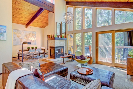 A modern living room with large windows, a leather sectional, a fireplace, and a dining area. Sunlight streams in through the windows, illuminating the space.