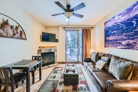 A cozy living room with a mounted TV, a fireplace, a brown leather sofa, a small dining table, and a wall art featuring a mountain scene. The room has a window with a view of a snowy landscape.