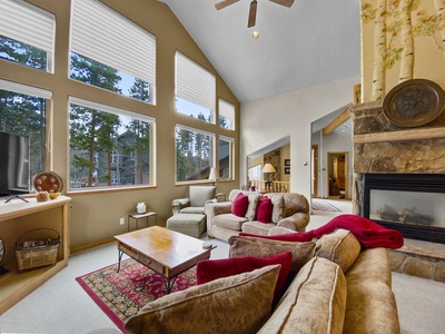 A cozy living room with large windows, a high ceiling, a fireplace, sofas with red pillows, and a wooden coffee table in a cabin-like setting.