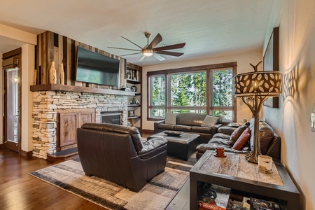 A cozy living room features a stone fireplace, a large window with a view of greenery, leather seating, a ceiling fan, and a patterned floor lamp.