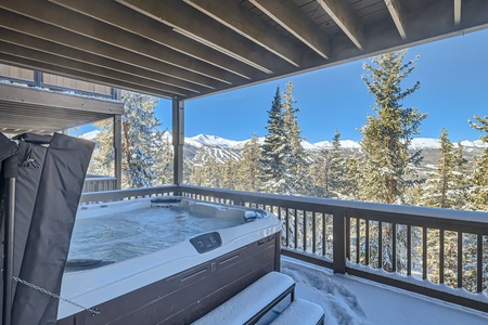 Outdoor hot tub on a snow-covered balcony with a view of pine trees and distant mountain peaks under a clear blue sky.