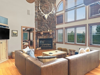 A spacious living room features a sectional sofa, a stone fireplace with mounted antlers above, large windows with blinds, and a wooden coffee table.