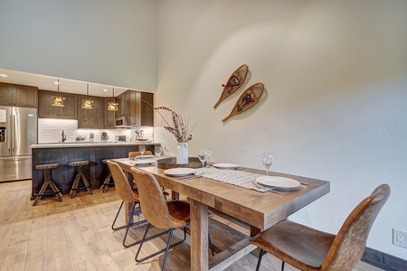 A kitchen and dining area with a wooden table, four chairs, place settings, and decorative snowshoes on the wall. The kitchen has bar stools, pendant lights, and stainless steel appliances.