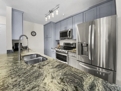 Modern kitchen with stainless steel appliances, granite countertops, and blue-gray cabinets. Features a dual sink, gas stove, microwave, and refrigerator under bright ceiling lights.