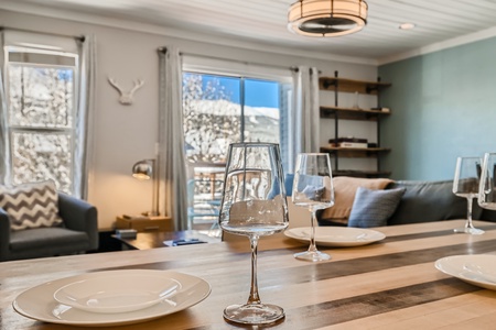 A modern dining area with a table set for a meal, featuring empty plates and wine glasses. Large windows in the background allow natural light to brighten the room, which includes a shelf and cozy seating.