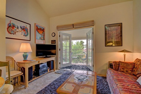 A cozy living room featuring a sofa, a coffee table, a TV console with a flat-screen TV, wall art, and an open double door leading to a porch with greenery outside.