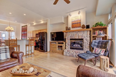 A cozy living room with a stone fireplace, armchair, and flat-screen TV. An open kitchen with barstools and a dining area with a chandelier are visible in the background.