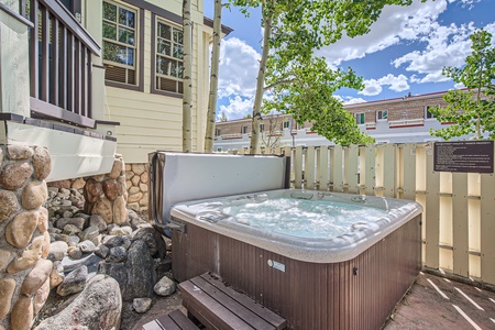 A hot tub on a stone patio next to a wooden fence and a beige house with large windows, under a partly cloudy sky.