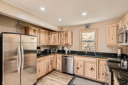 A modern kitchen with wooden cabinets, stainless steel appliances, granite countertops, and a window above the sink. The room is well-lit with recessed lighting.