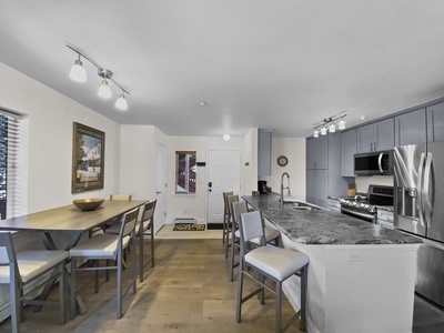 A modern kitchen with a marble countertop island, stainless steel appliances, and adjacent dining area with a wooden table and chairs. White walls and light wooden flooring are present throughout.