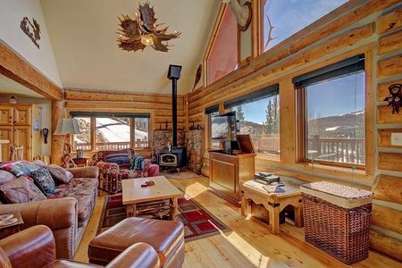 A cozy log cabin interior featuring a wood stove, large windows with scenic views, and rustic decor. The room includes a comfortable seating area, a TV, and natural wood furniture.