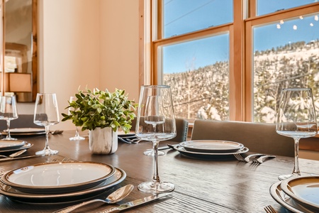 A dining table set with plates, wine glasses, cutlery, and a potted plant, positioned near a large window with a snowy mountain view in the background.