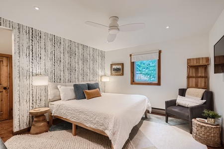 A bedroom with a king-sized bed, bedside tables with lamps, an armchair, a window, and a wall-mounted TV. The room features a nature-themed accent wall and a ceiling fan.