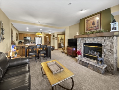 A cozy living room with a stone fireplace, leather sofa, wooden coffee table, and a dining area in the background. The room is decorated with rustic elements.