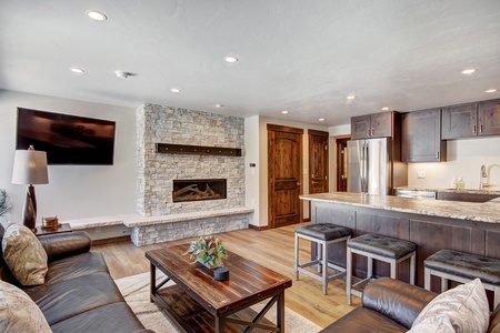 Modern living room and kitchen with a stone fireplace, mounted TV, wooden cabinetry, stainless steel refrigerator, granite countertops, and a seating area with dark furniture and a small coffee table.