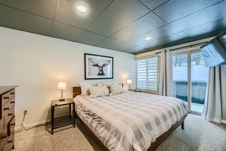 A bedroom with a double bed, checkered bedding, two bedside tables with lamps, a deer print on the wall, a dresser, and a large window with a view of snow outside.